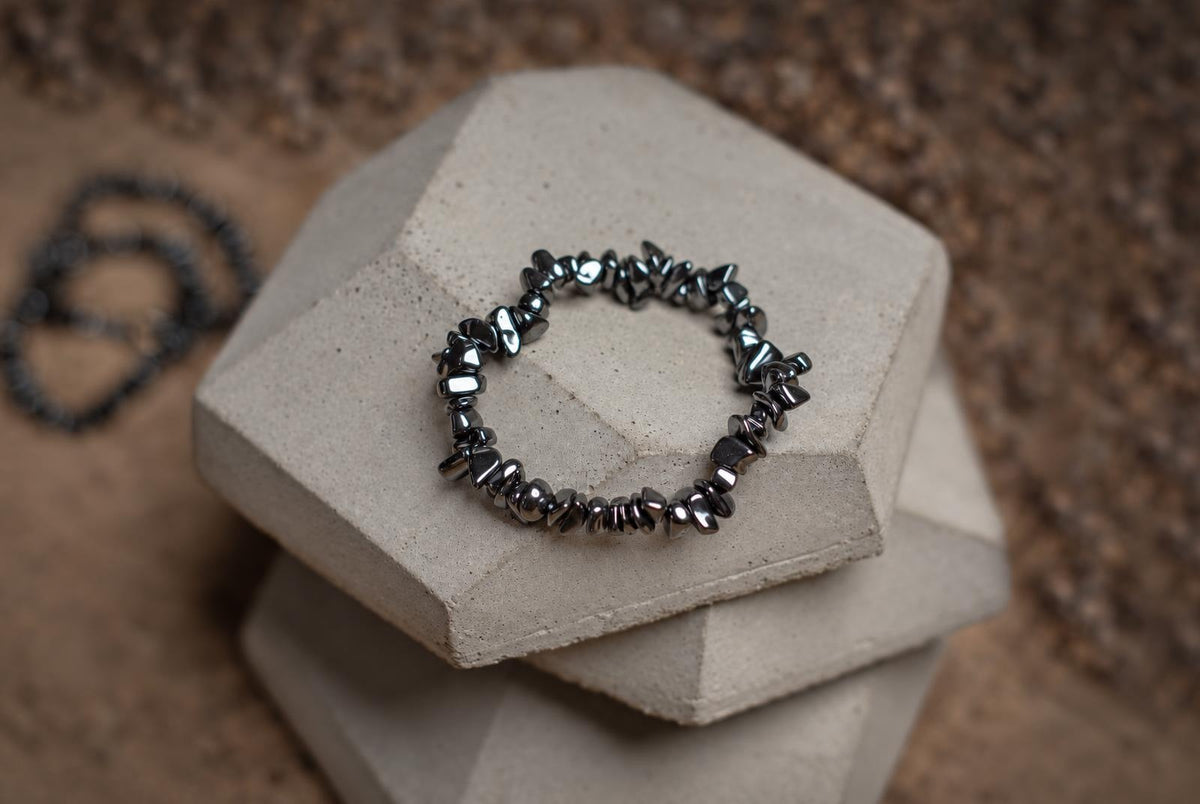 A bracelet of dark metallic-looking chip beads on stacked grey blocks, with blurred bracelets in the background.