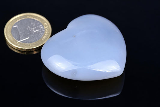 A polished pale purple agate heart rests beside a coin on a dark reflective surface.