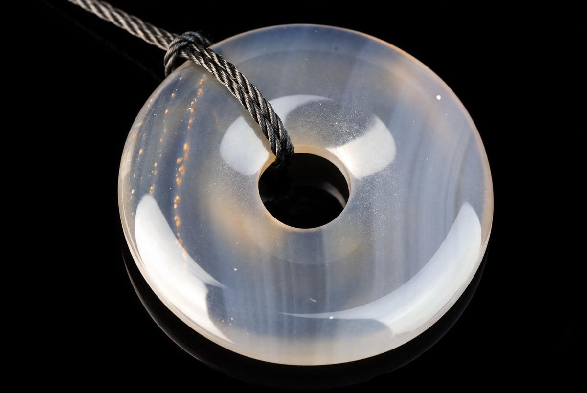 A translucent, donut-shaped pendant with a central hole and subtle internal banding, suspended by a twisted cord against a dark surface.