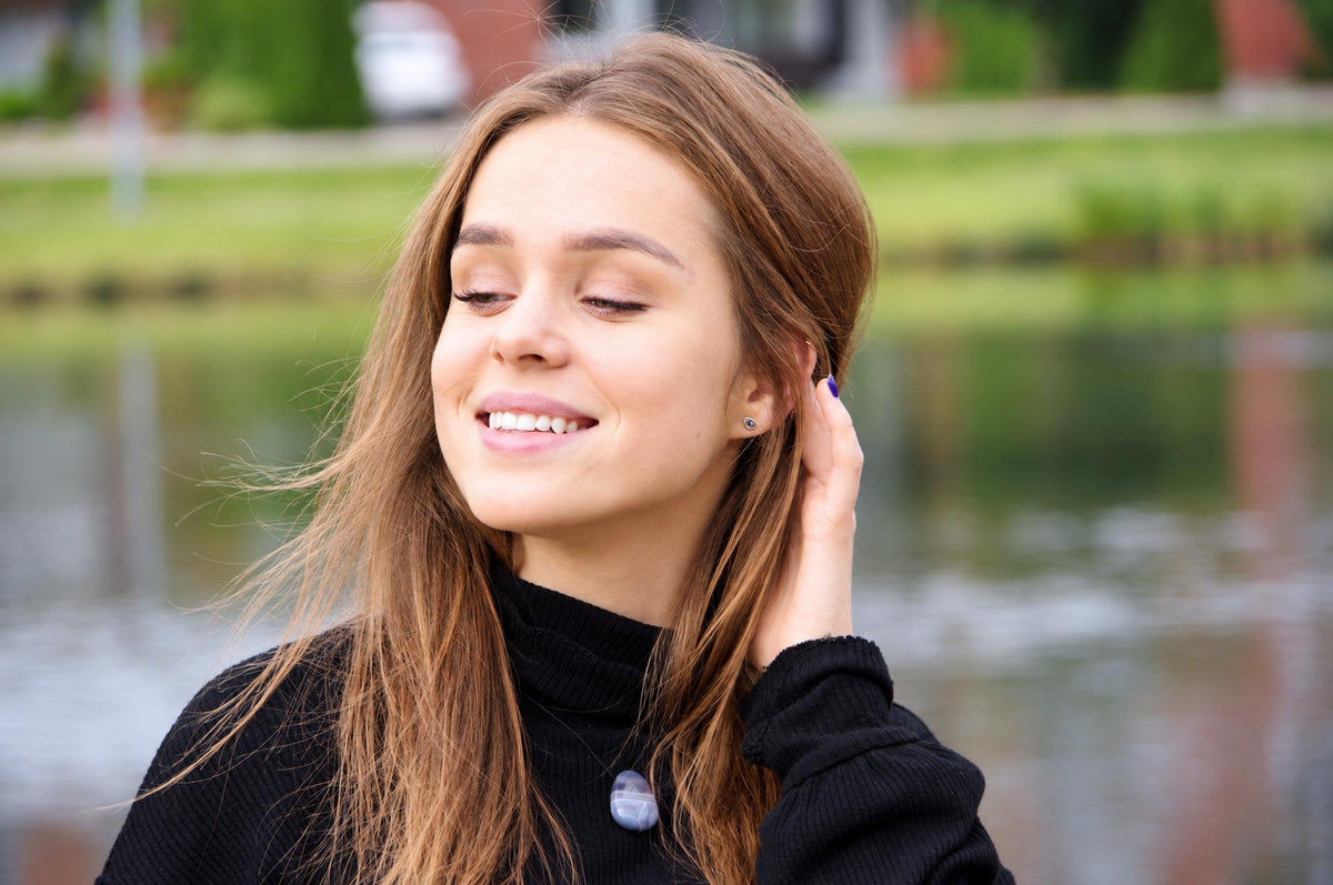 A smiling woman with long brown hair wears a black turtleneck and a round, banded blue-grey agate pendant on a dark surface.