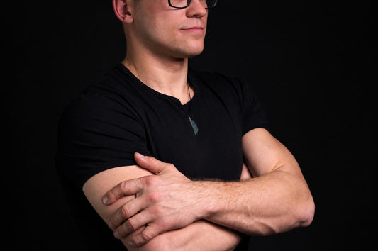 A man in a black t-shirt wears a dark green, wing-shaped pendant on a thin cord against a dark background.