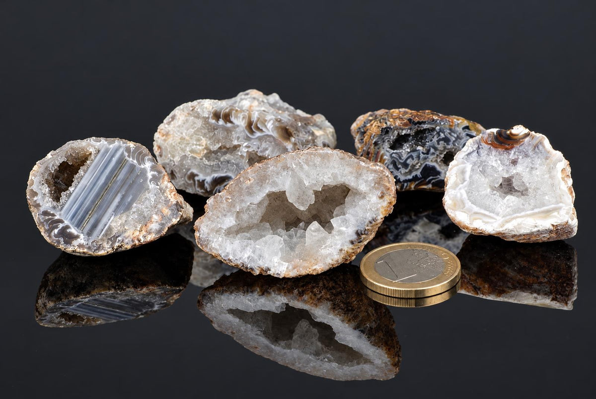 Open agate pieces show pale crystal cavities and grey banding beside a coin, with their reflections visible on a glossy dark surface.