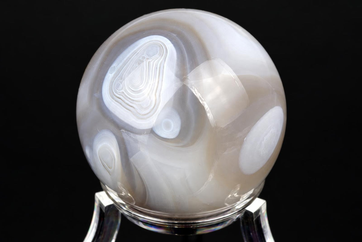 A polished agate sphere with swirling white and grey bands sits on a clear stand against a black background.