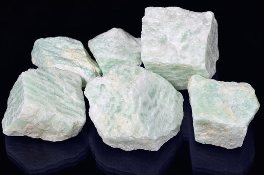 Six raw Amazonite India mineral specimens with pale green and white crystalline textures on a dark reflective surface.