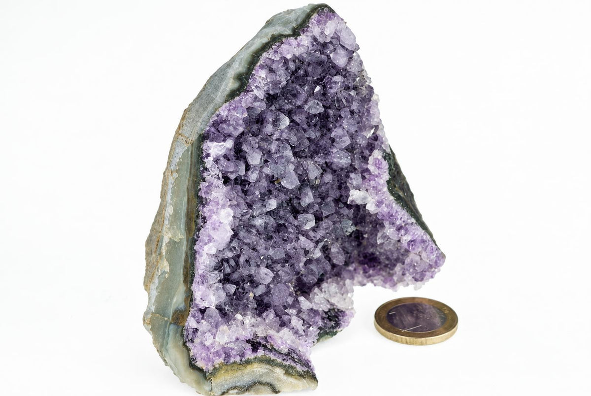 A cut amethyst geode with purple crystals inside a grey-green outer shell, next to a coin on a white surface.
