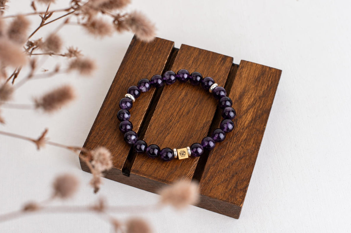 A bracelet with round purple beads and a gold-toned square charm rests on a dark wooden surface.