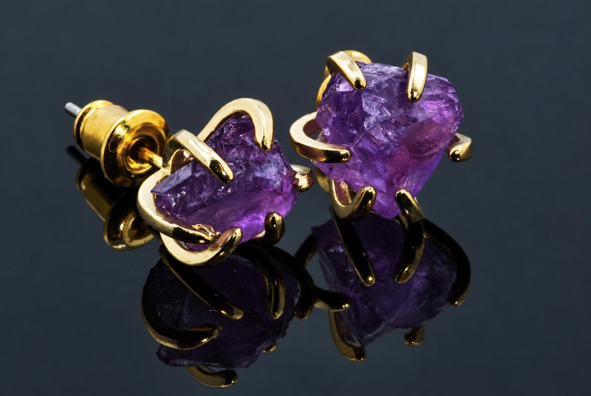 A pair of gold-toned stud earrings with rough-cut purple amethyst stones held by curved prongs, displayed on a dark reflective surface.