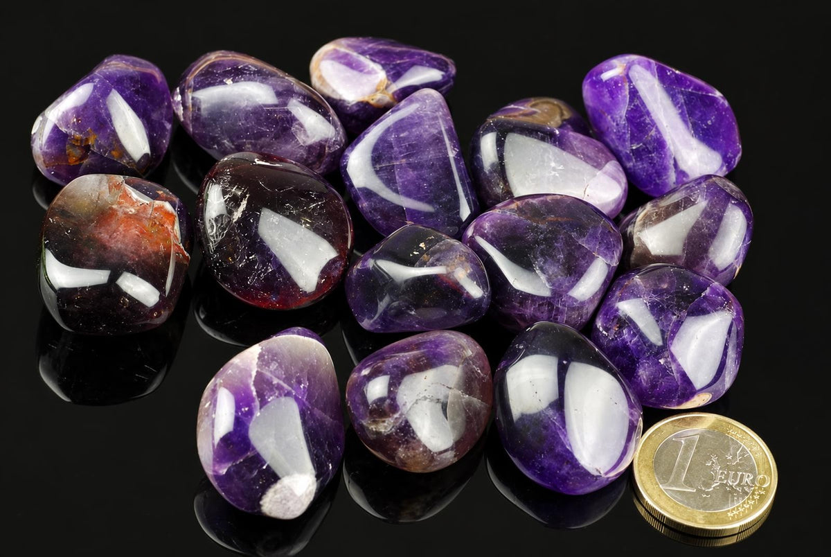 A pile of polished purple amethyst tumbled stones with white inclusions on a dark surface, next to a one euro coin.