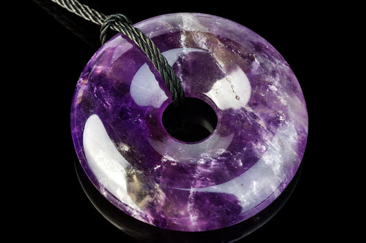 A polished purple and white donut-shaped pendant with a central hole, threaded on a dark cord against a black background.