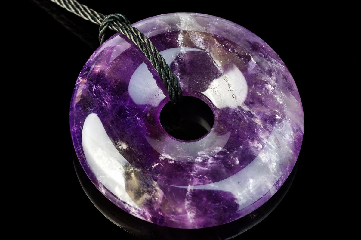 A polished purple and white donut-shaped pendant with a central hole, threaded on a dark cord against a black background.