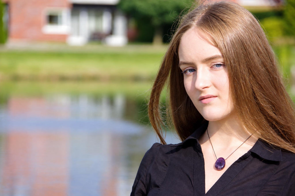 A woman wears a black shirt and a necklace with a dark purple oval pendant on a thin cord, outdoors near water.