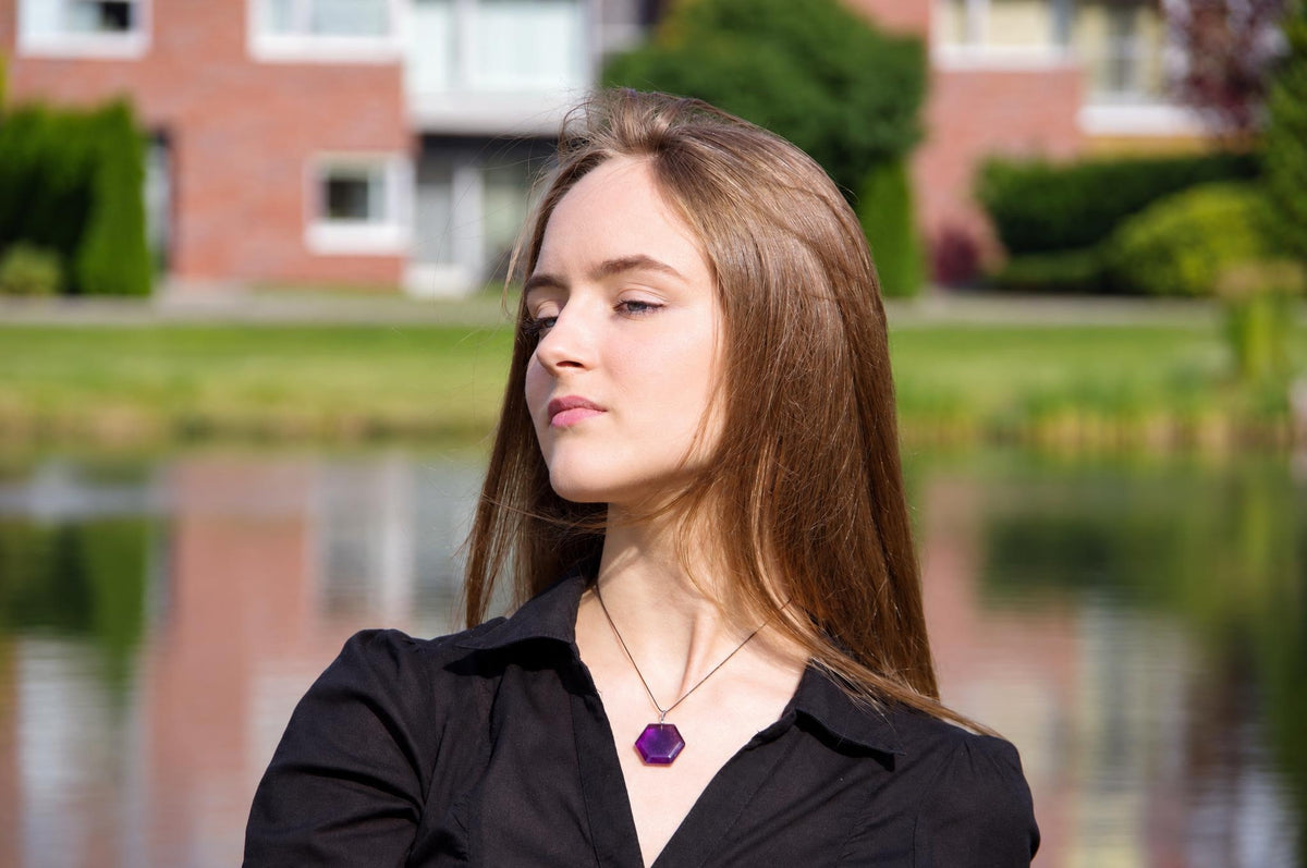 A woman wears a necklace with a purple hexagonal pendant on a thin chain, against a blurred outdoor background.