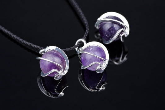 Three silver-toned wire pendants with purple spheres on a black cord, resting on a dark reflective surface.
