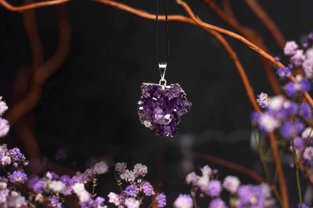 A silver-toned pendant with a rough, purple amethyst crystal cluster hangs from a black cord against a dark background with blurred branches and flowers.