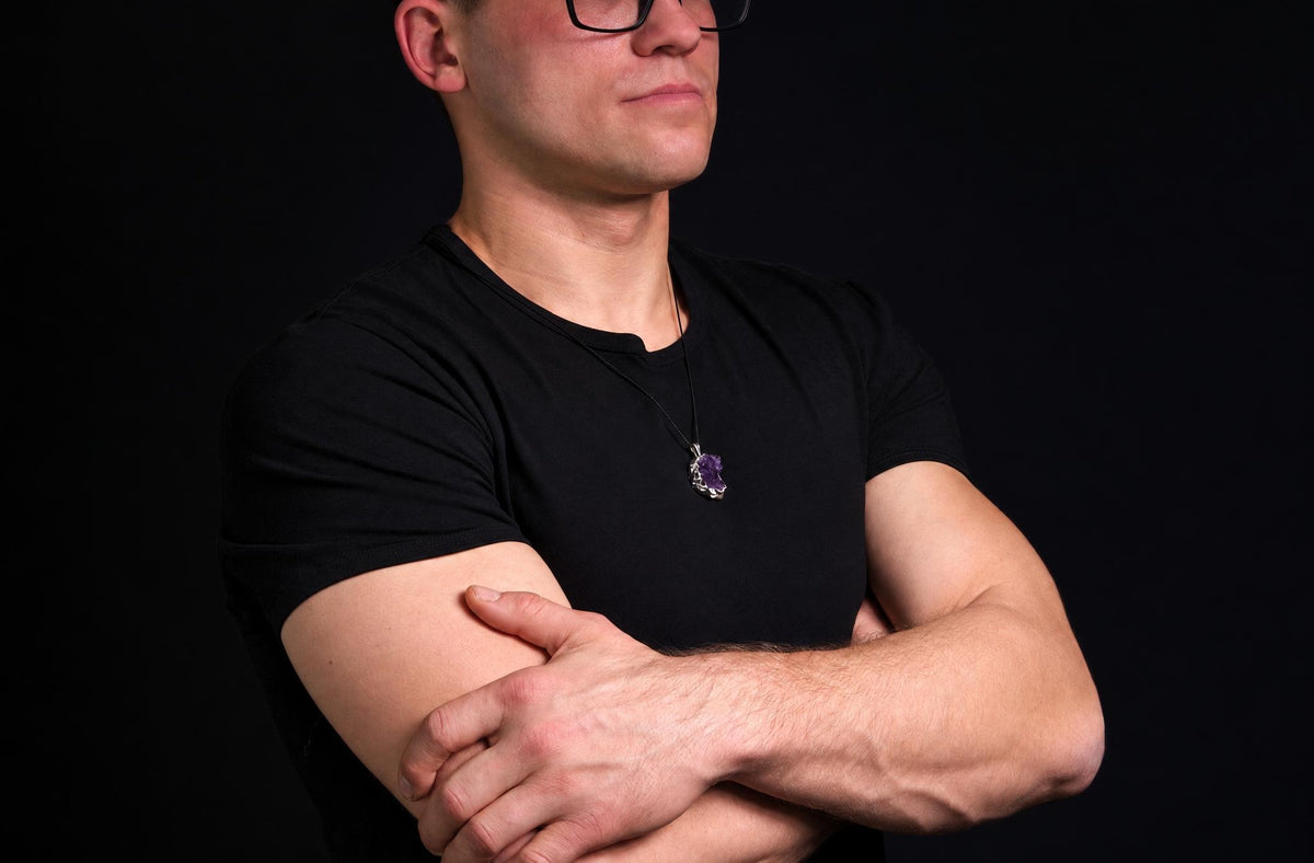 A man in a black t-shirt wears a pendant with a purple amethyst stone on a dark surface.
