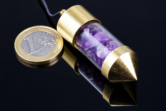 A brass pendulum with a pointed tip and clear tube filled with purple amethyst crystals, next to a one-euro coin on a dark surface.
