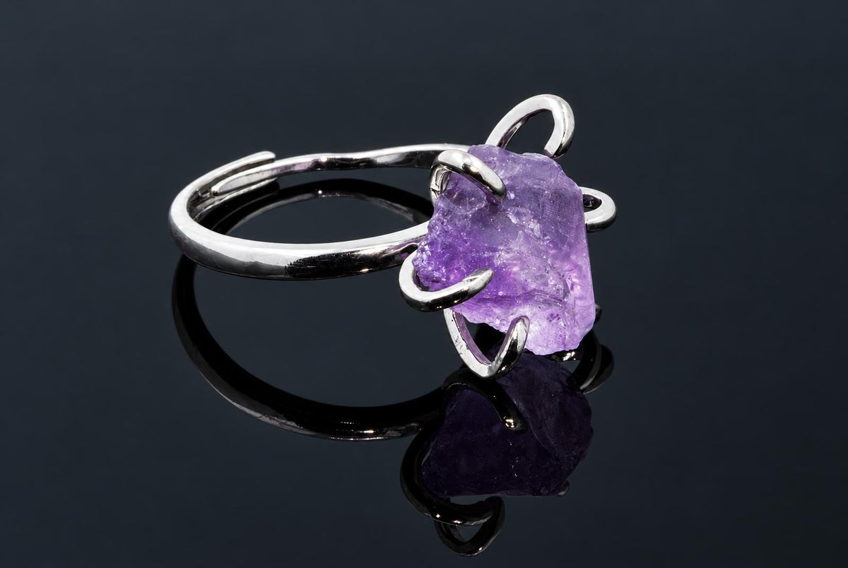 A silver-tone ring holds a rough purple crystal in curved arms on a dark reflective surface.