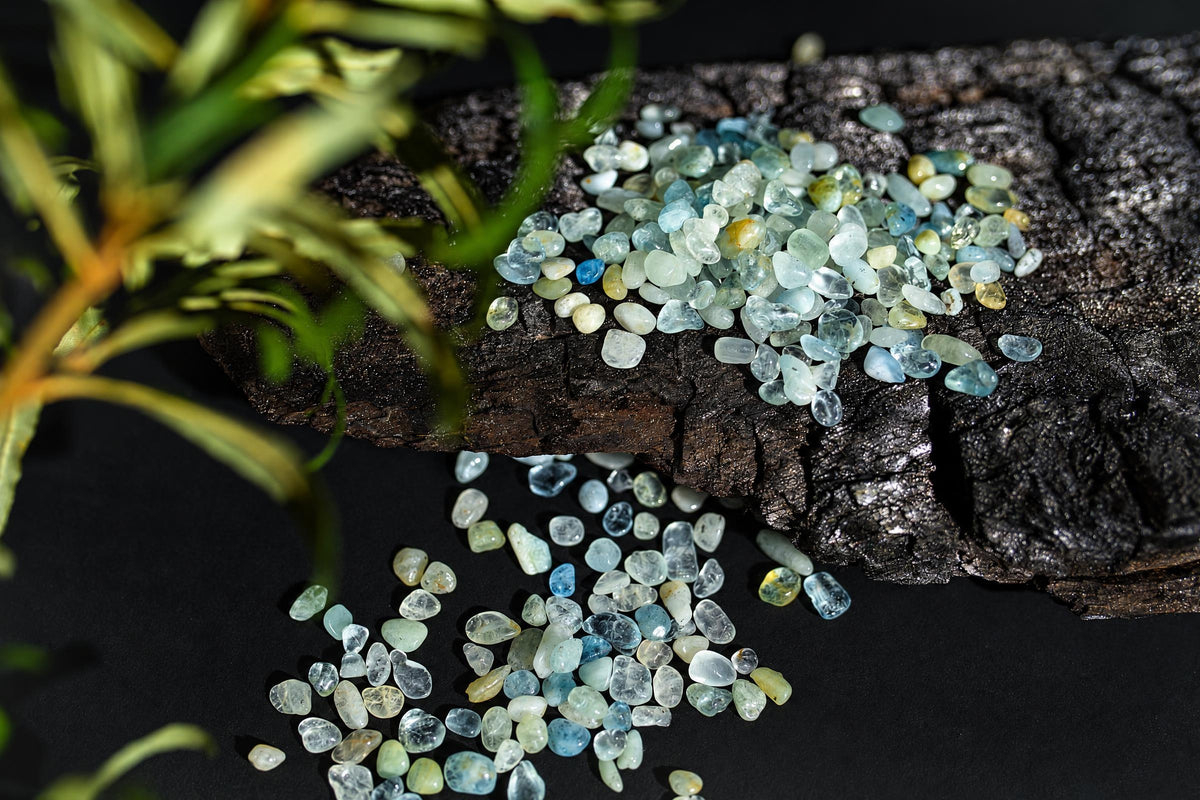 Small translucent pale blue and yellowish stones are scattered on a dark textured surface beneath green foliage.