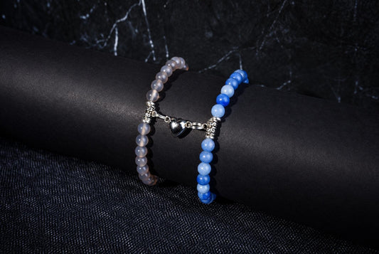 Two beaded bracelets on a dark surface: one with pale beads and a silver heart charm, the other with blue beads.