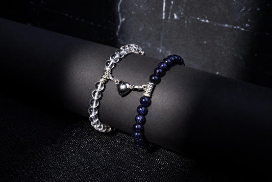 Two beaded bracelets on a dark surface: one with clear beads and a silver heart charm, the other with deep blue beads.