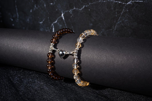Two beaded bracelets on a dark cylindrical form: one with brown beads, the other with clear and yellow beads. A silver heart charm connects them.