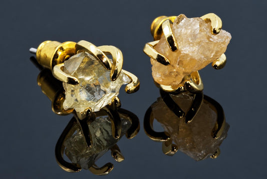 A pair of gold-coloured stud earrings with irregular, rough-cut citrine stones held by curved metal claws on a dark reflective surface.