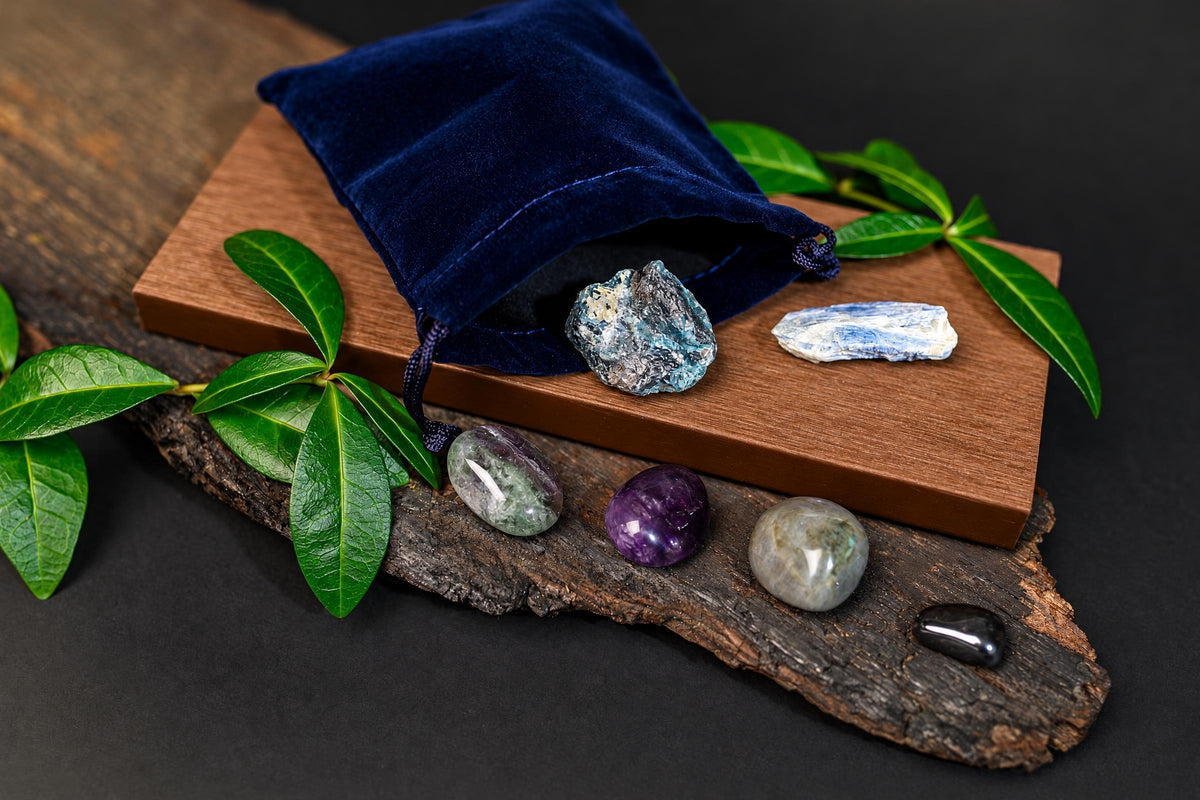 A blue velvet pouch on a wooden board with green leaves, holding rough blue crystals. Polished stones in purple, grey, and black rest on a dark wood slice below.