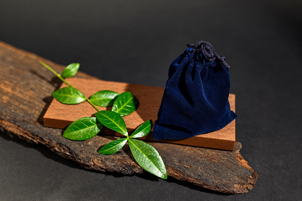 A dark blue velvet pouch with a drawstring sits on a wooden block next to green leaves on a rustic wood slab.