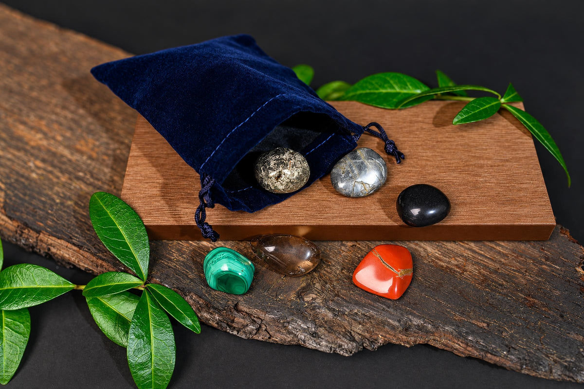 An open dark blue drawstring pouch and colourful polished stones are arranged on wooden surfaces beside green leaves.