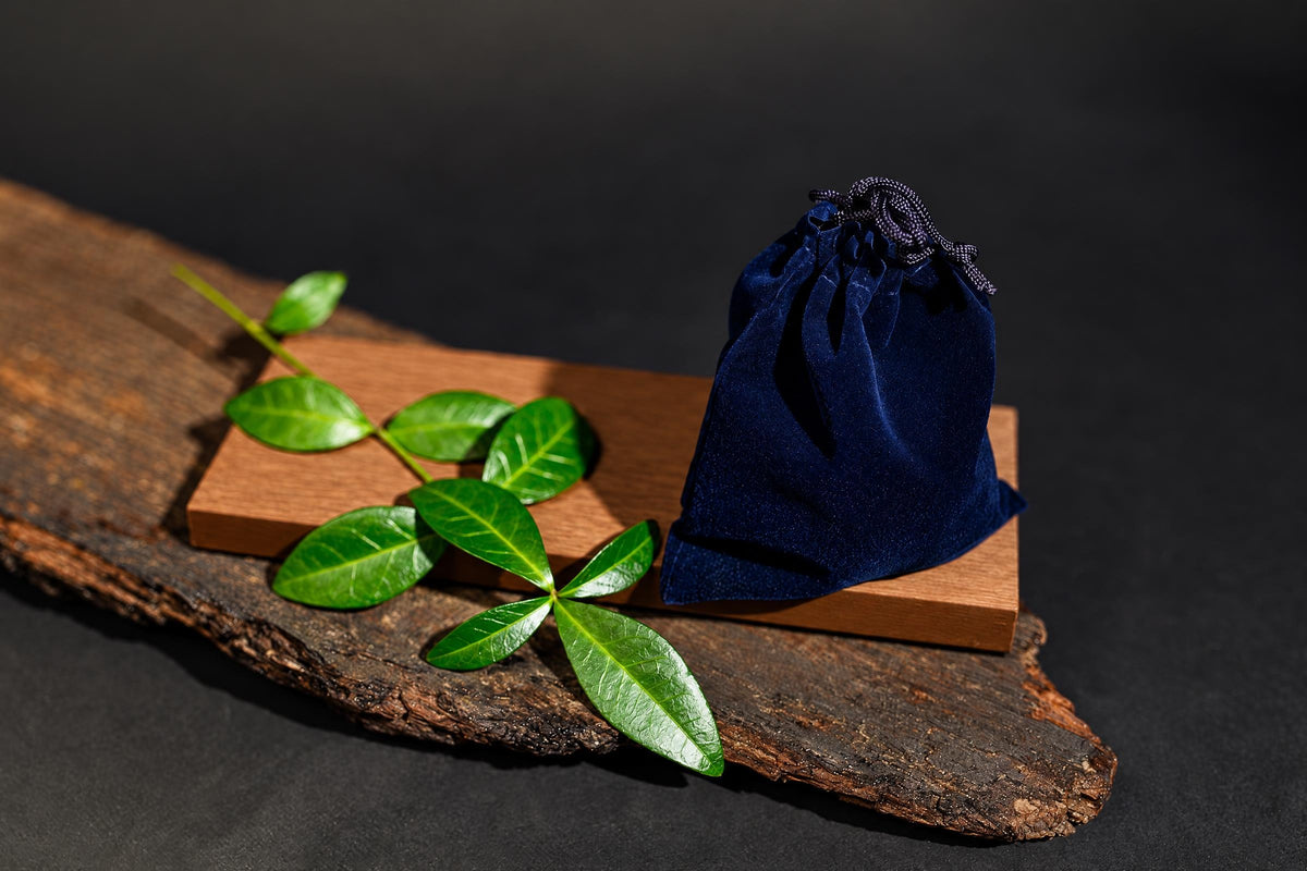 A dark blue velvet pouch with a drawstring sits on a wooden block beside green leaves on a rustic wood slab.