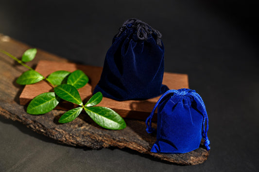 Two blue velvet drawstring pouches sit on a wooden surface with green leaves nearby.