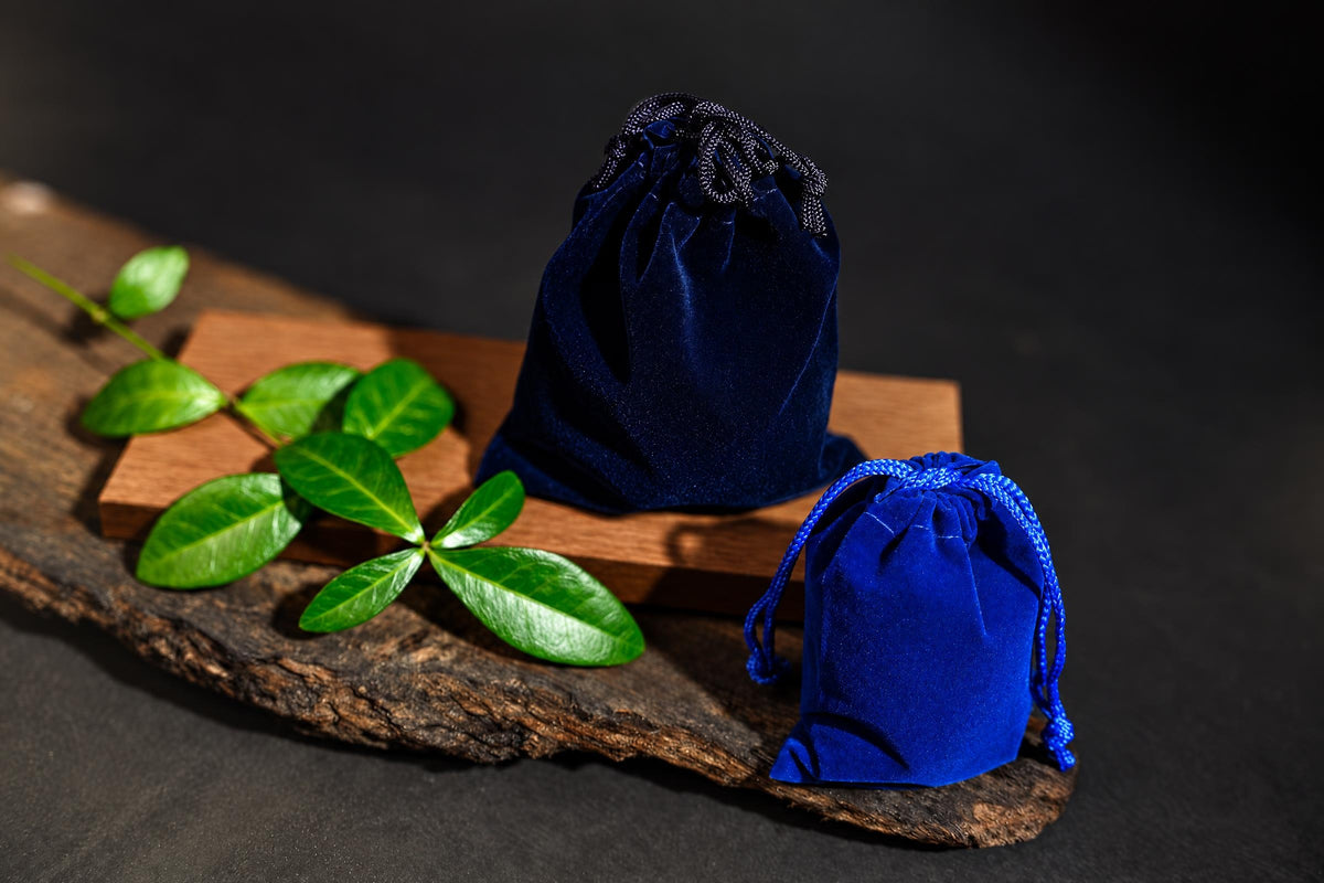 Navy-blue and bright blue drawstring pouches rest on a wooden surface beside green leaves against a dark background.