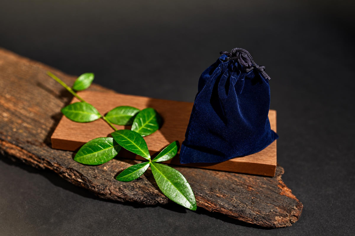 A dark blue velvet pouch with a drawstring sits on a wooden block, next to green leaves on a rustic wood slab.