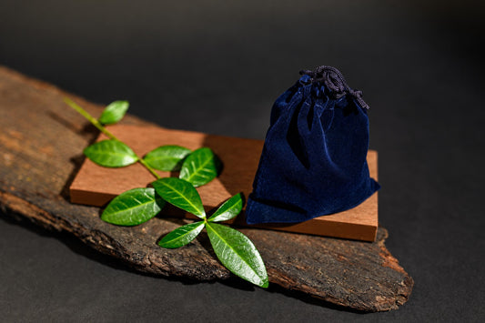 A dark blue velvet pouch with a drawstring sits on a wooden block, next to green leaves on a rustic wood slab.