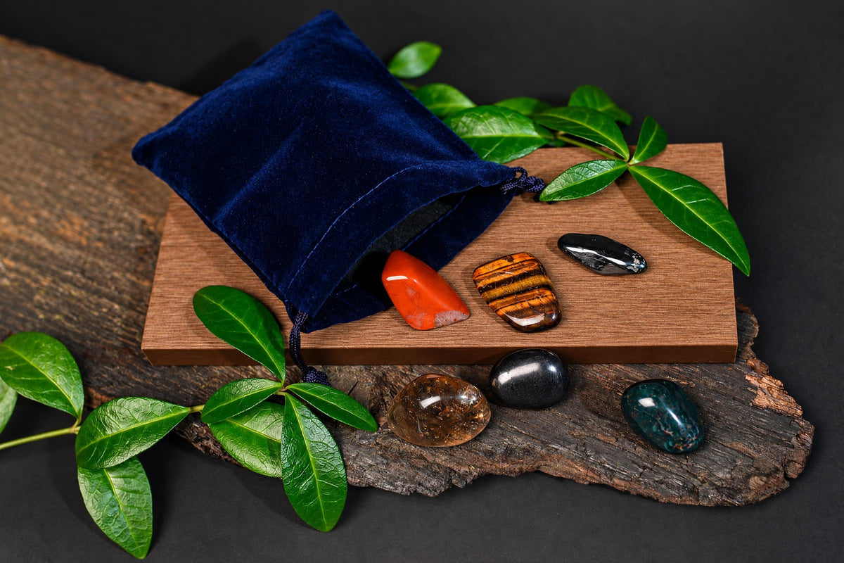 A blue velvet pouch spills polished stones onto a wooden board and dark surface, surrounded by green leaves.