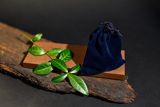 A dark blue velvet pouch with a drawstring sits on a wooden block next to green leaves on a rustic wood slab.