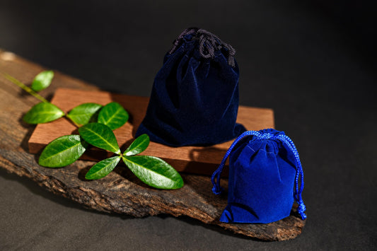 Two blue velvet drawstring pouches sit on a wooden surface with green leaves nearby.