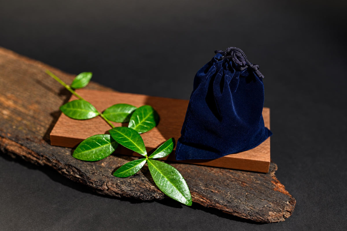 A dark blue velvet pouch with a drawstring sits on a wooden block, next to green leaves on a rustic wood slab.