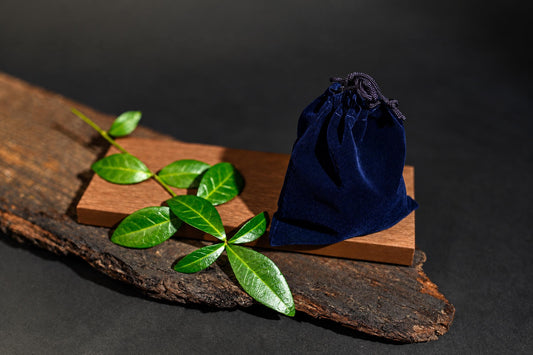 A dark blue velvet pouch with a drawstring sits on a wooden block next to green leaves on a rustic wood slab.