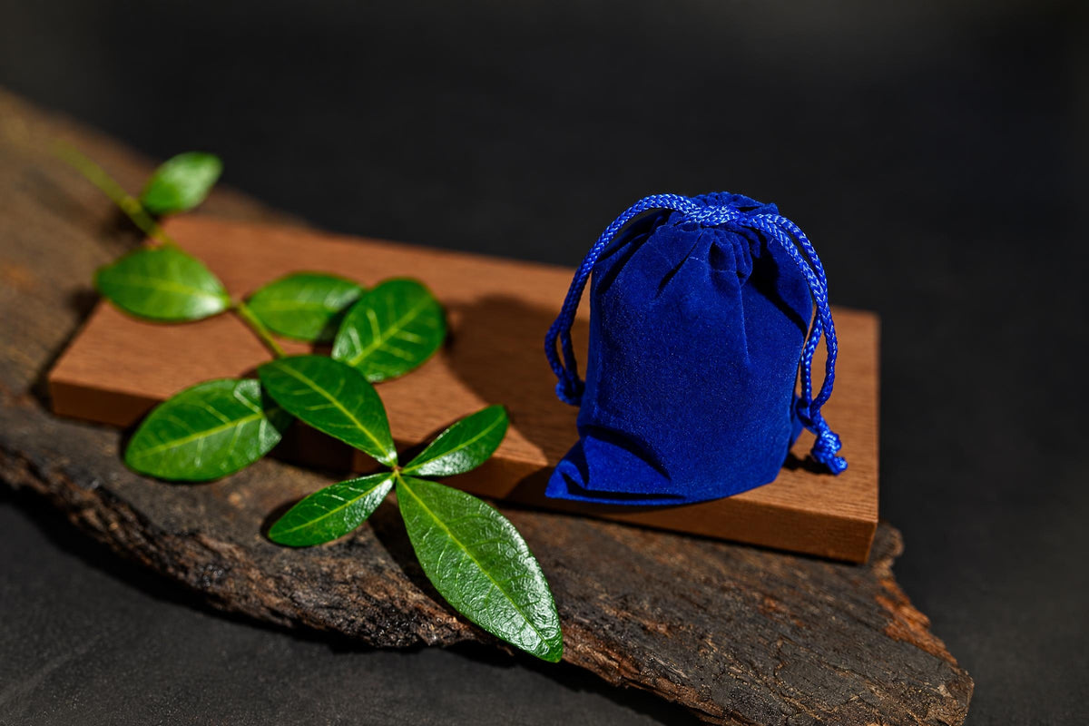 A blue velvet pouch with a drawstring sits on a wooden block next to green leaves on a dark surface.