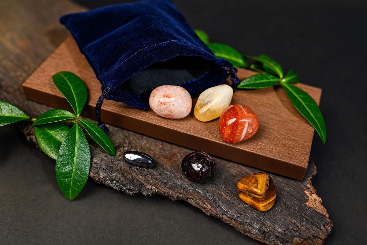 A blue velvet pouch with a drawstring sits on a wooden board, surrounded by polished stones in pink, yellow, orange, black, and brown tones.