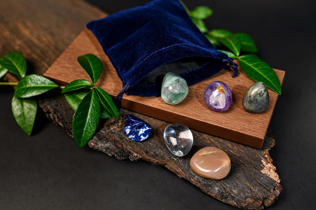 An open blue pouch rests beside polished stones in varied colours on wooden surfaces with green leaves.