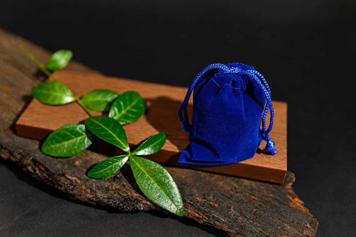 A blue drawstring pouch stands on a wooden board beside green leaves against a dark background.