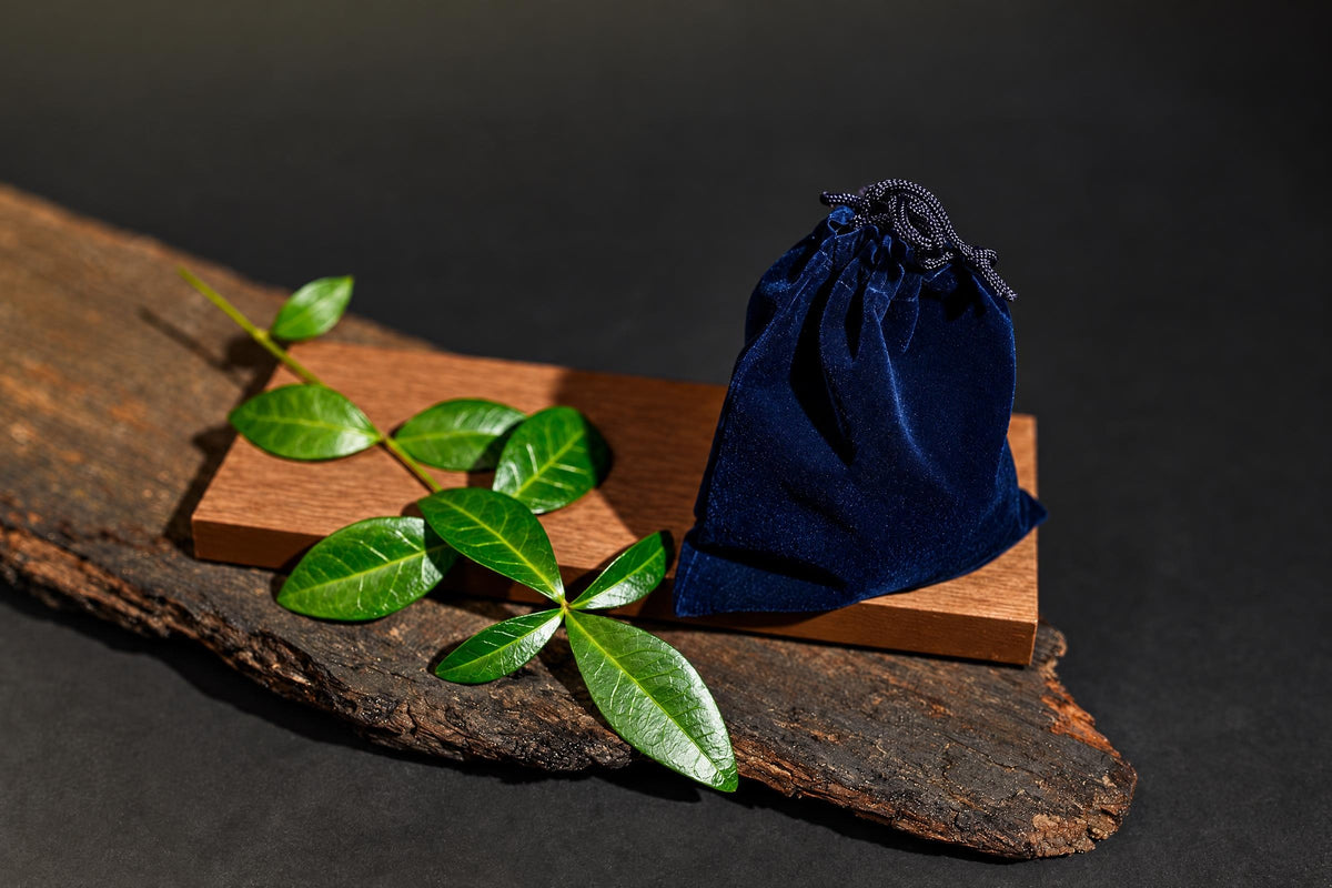 A dark blue velvet pouch with a drawstring sits on a wooden block next to green leaves on a rustic wood surface.