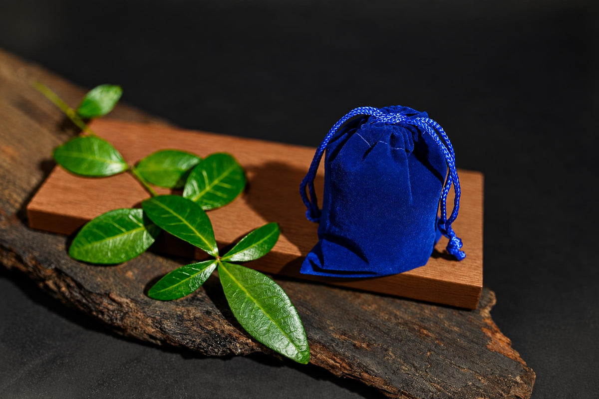 A blue velvet drawstring pouch sits on a wooden board with green leaves, all on a dark surface.