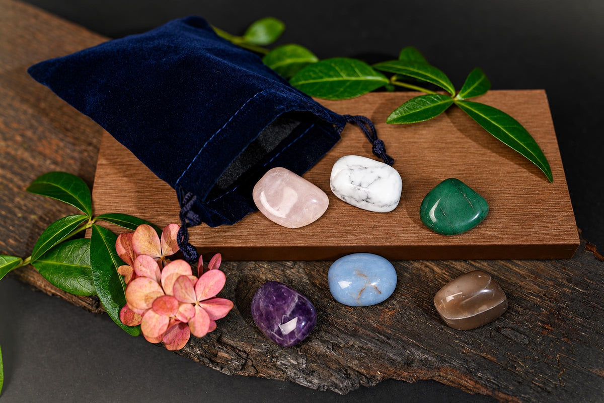 A dark blue velvet pouch with a drawstring sits on wood next to six polished stones in pink, white, green, purple, blue and brown.