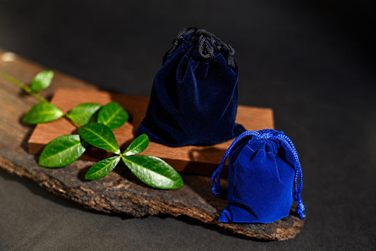 Two blue velvet drawstring pouches sit on a wooden surface with green leaves nearby.