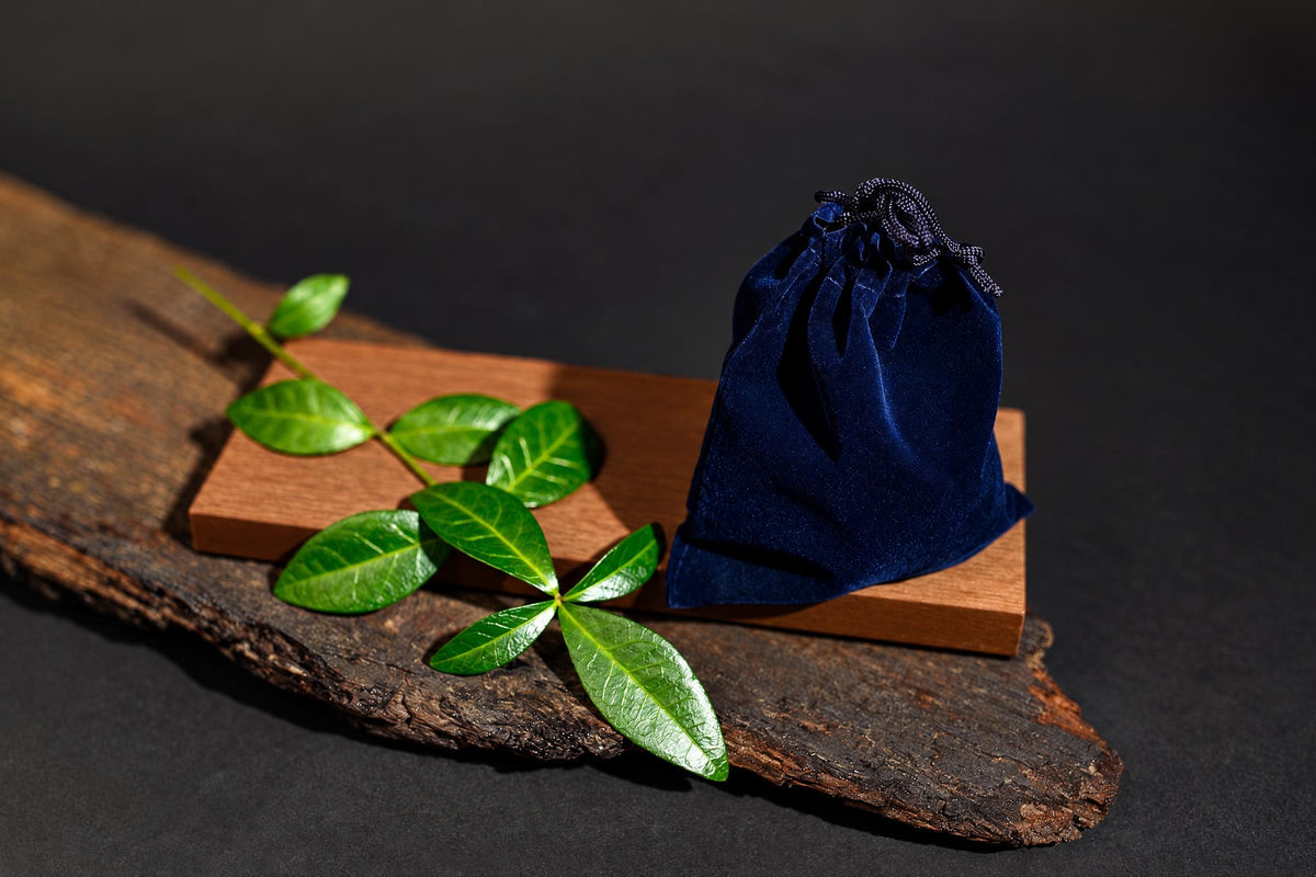 A dark blue velvet pouch with a drawstring sits on a wooden block next to green leaves on a rustic wood slab.