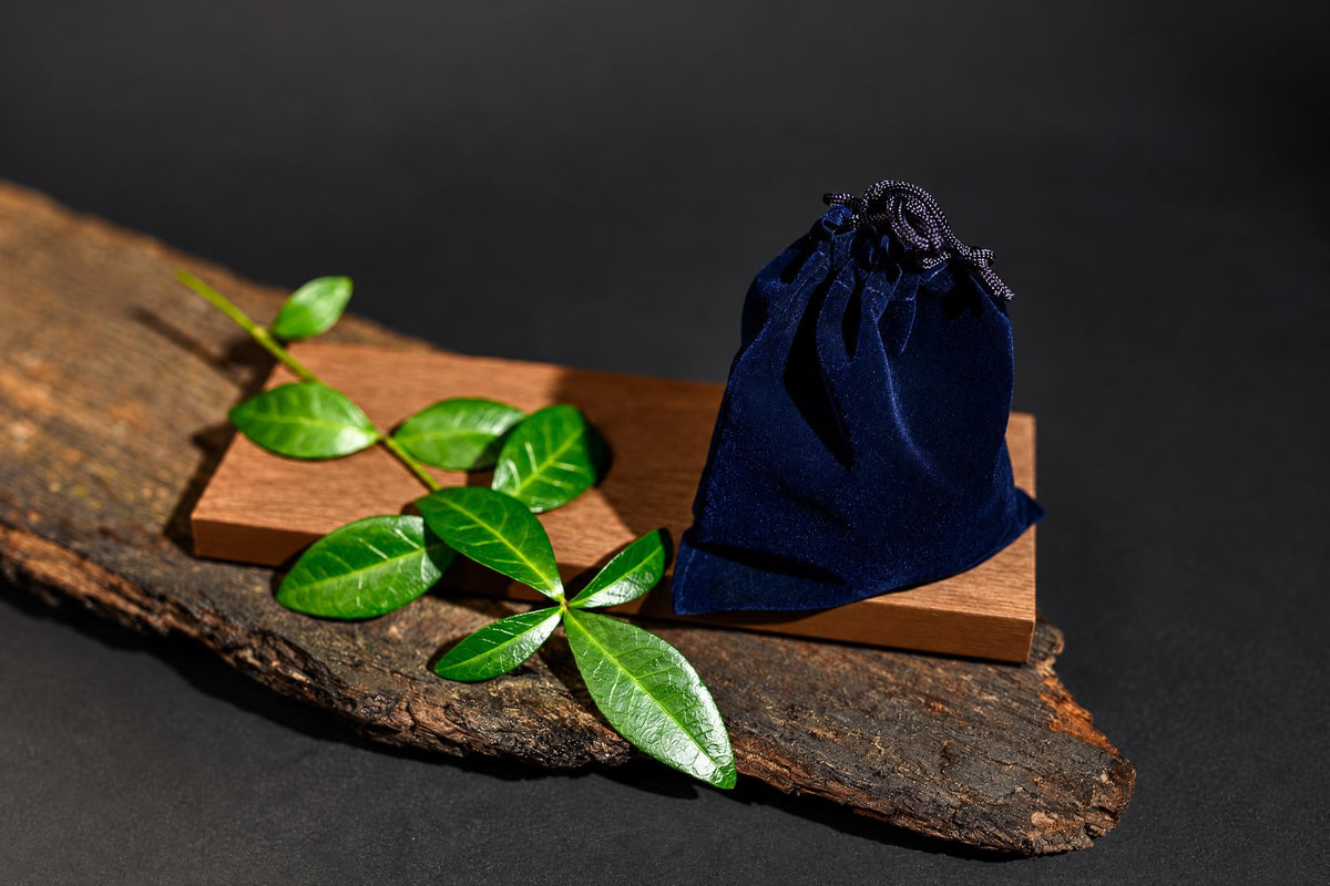 A dark blue velvet pouch with a drawstring sits on a wooden block, next to green leaves on a rustic wood slab.
