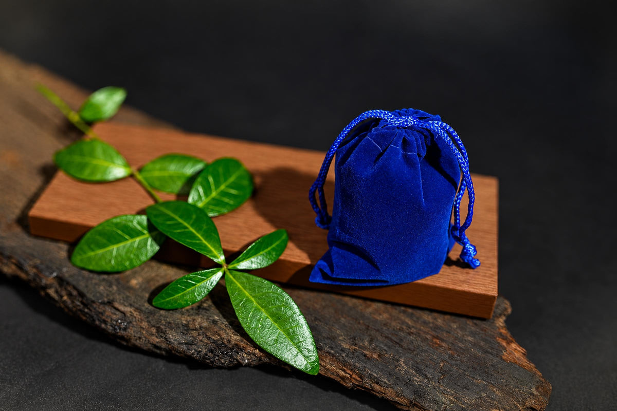 A blue velvet drawstring pouch sits on a wooden board with green leaves, all on a dark surface.